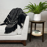 Gray sofa with a black and white patterned throw blanket, next to a wooden side table with a plant and books.